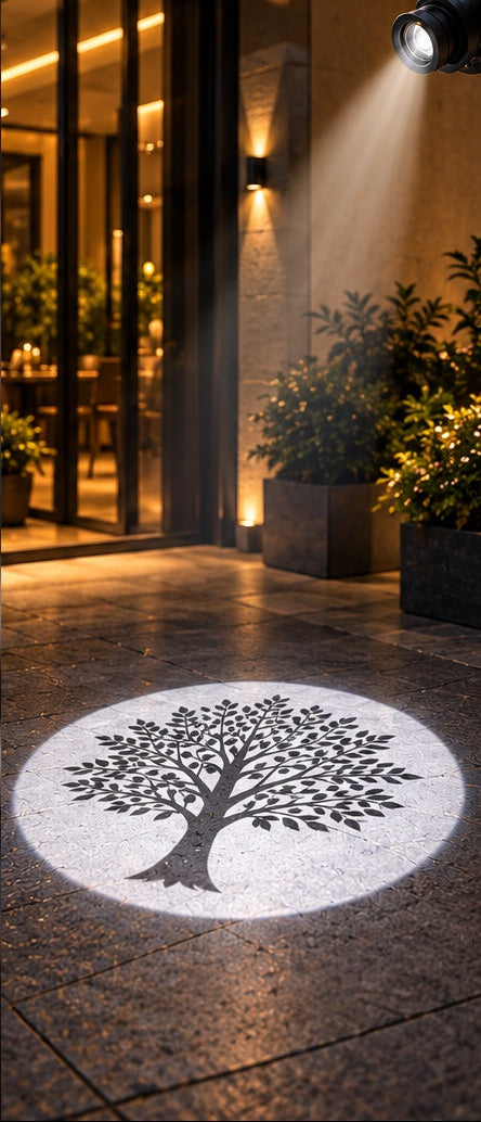 A black tree silhouette projected onto a stone patio floor in a circular white light beam, with a modern building entrance, plants, and warm wall lighting in the background.